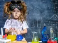 Mała chemik z rozczochranymi włosami i okularami ochronnymi, trzymająca kolby z kolorowymi płynami. W tle tablica z wzorami, idealna do eksperymentów chemicznych dla dzieci.