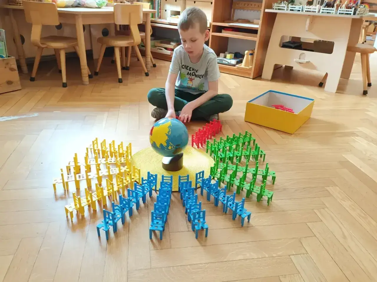 Chłopiec bawi się klockami-krzesłami wokół globusa, ucząc się przez zabawę. To przykład metody Montessori na czym polega nauka przez doświadczenie.