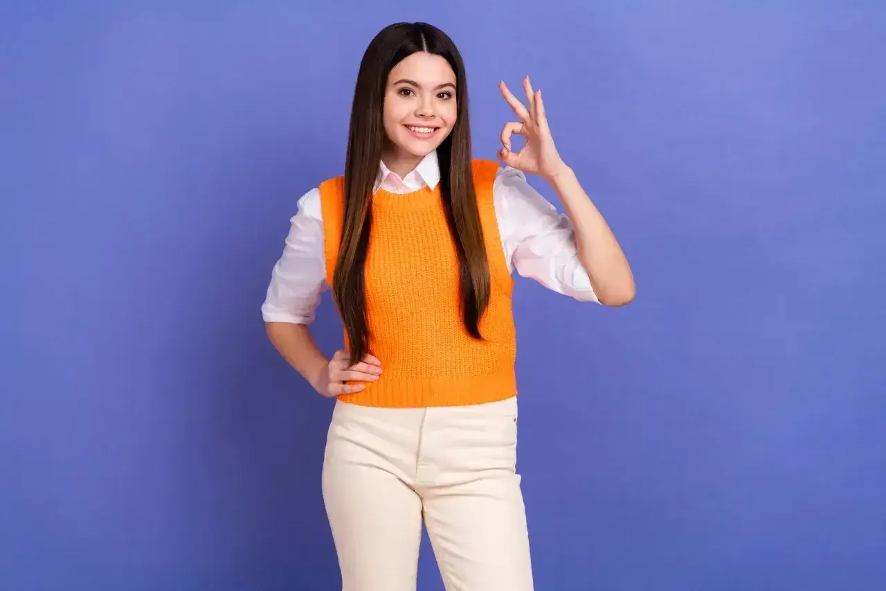 Uśmiechnięta dziewczyna w pomarańczowym sweterku pokazuje znak "OK". Czy matura ustna liczy się na studia? Tak, to ważny krok!