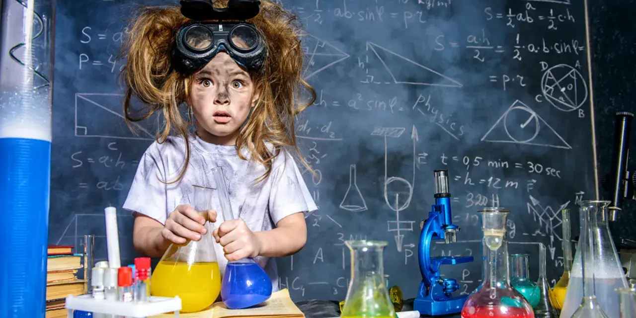 Mała chemik z rozczochranymi włosami i okularami ochronnymi, trzymająca kolby z kolorowymi płynami. W tle tablica z wzorami, idealna do eksperymentów chemicznych dla dzieci.