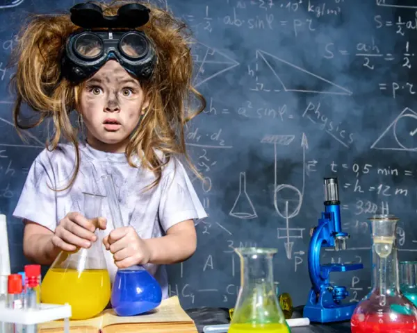 Mała chemik z rozczochranymi włosami i okularami ochronnymi, trzymająca kolby z kolorowymi płynami. W tle tablica z wzorami, idealna do eksperymentów chemicznych dla dzieci.