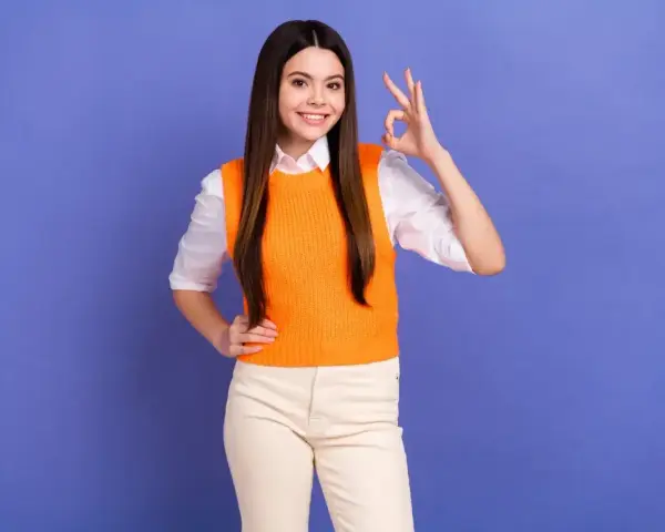 Uśmiechnięta dziewczyna w pomarańczowym sweterku pokazuje znak "OK". Czy matura ustna liczy się na studia? Tak, to ważny krok!