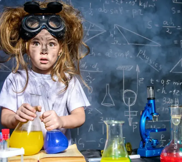 Mała chemik z rozczochranymi włosami i okularami ochronnymi, trzymająca kolby z kolorowymi płynami. W tle tablica z wzorami, idealna do eksperymentów chemicznych dla dzieci.