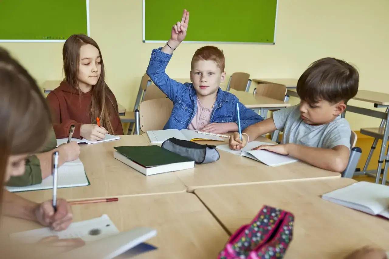 Dzieci w klasie piszą w zeszytach, jeden chłopiec podnosi rękę.