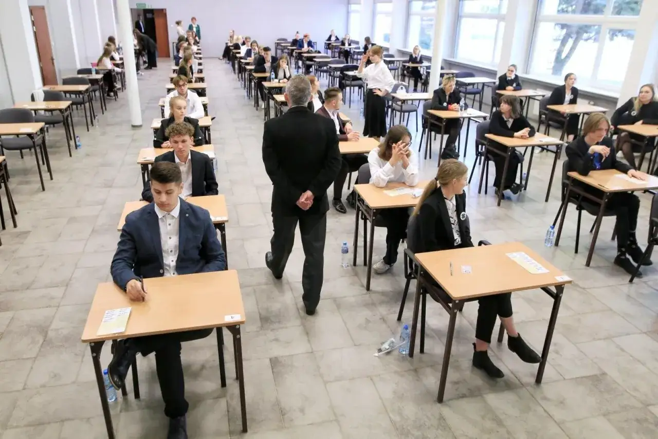 Uczniowie siedzą w ławkach podczas egzaminu. Czy matura próbna jest obowiązkowa? Nauczyciel nadzoruje salę.